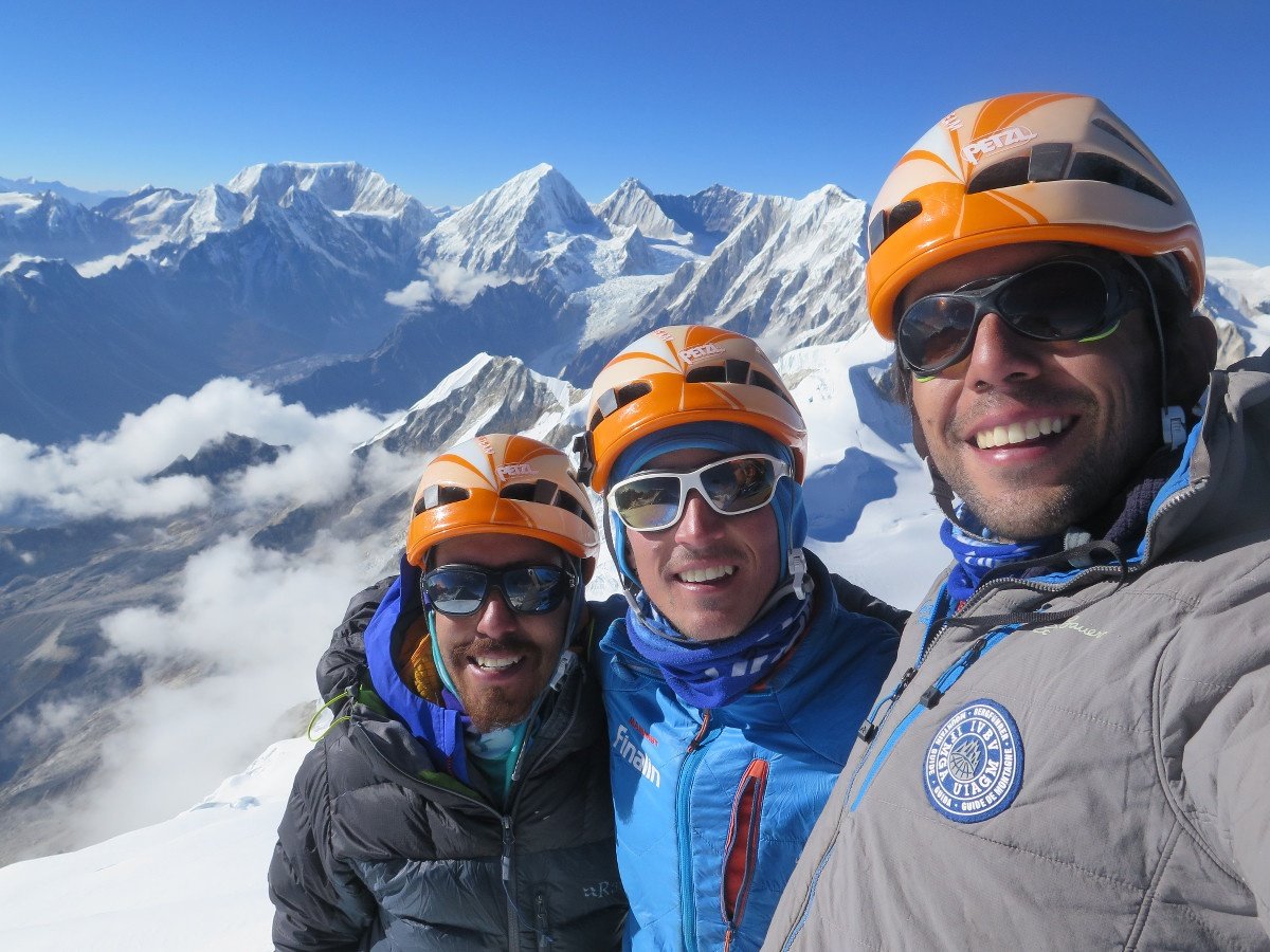 three smiling climbers wearing orange and white helmets at the summit of a mountain with snowy peaks beyond