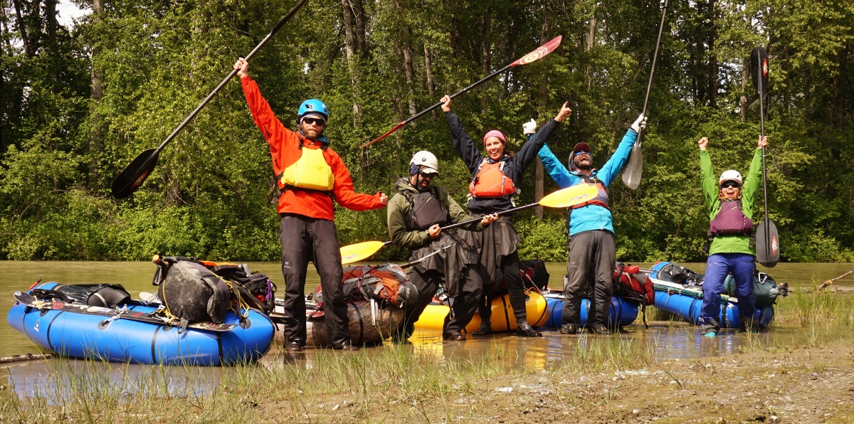 Five adventurers smile and raise their arms and paddles in the air while standing next to their packrafts
