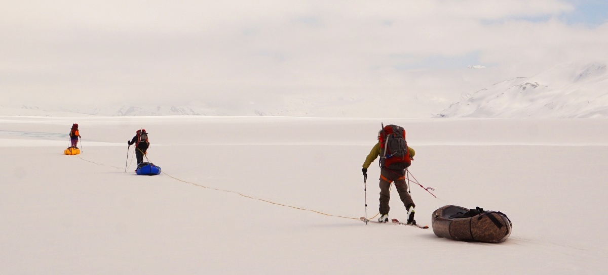 Team of three walk across a snow flat using trekking poles and dragging their packrafts like sleds