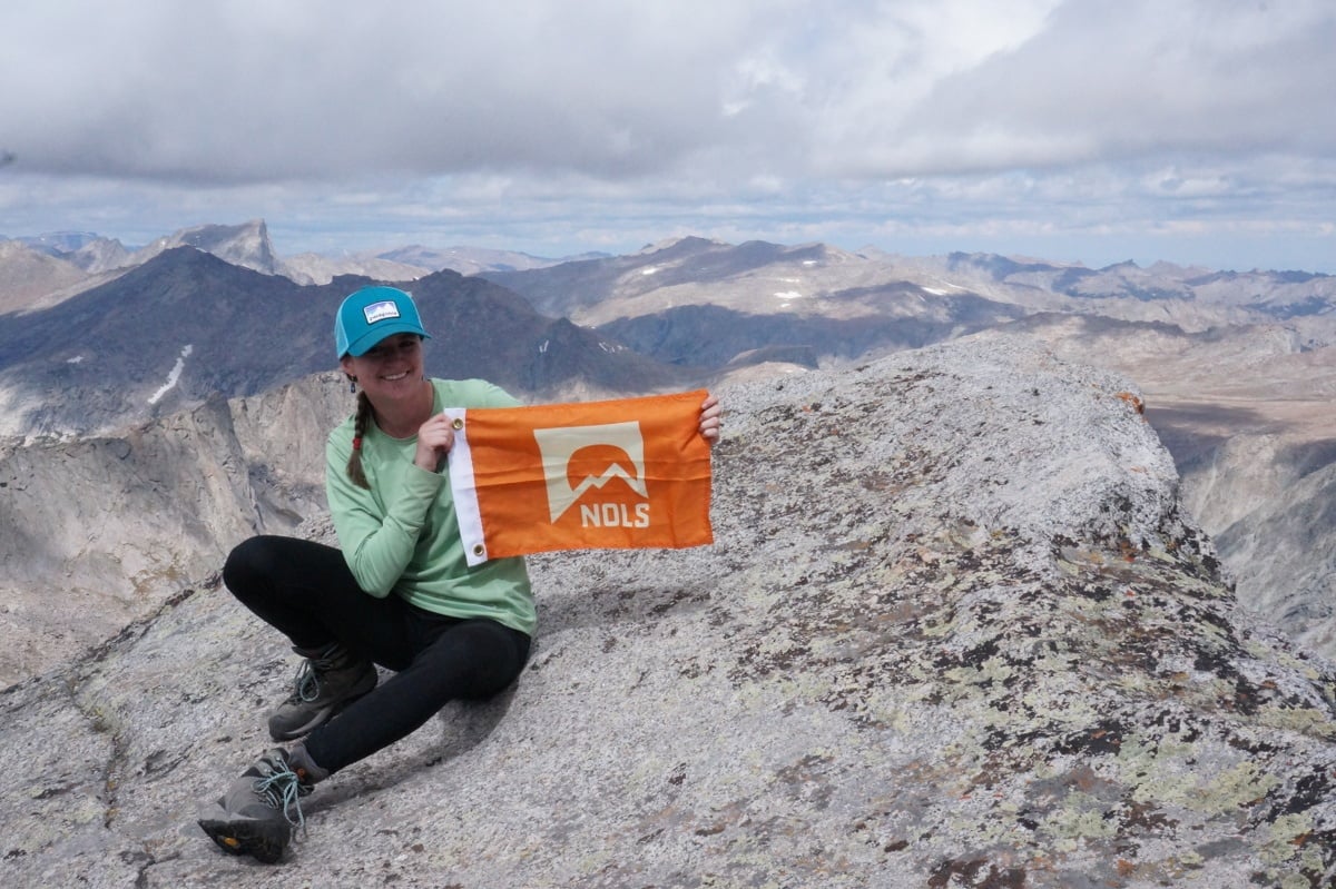 Maddy smiling and holding an orange NOLS summit flag on a peak in the mountains