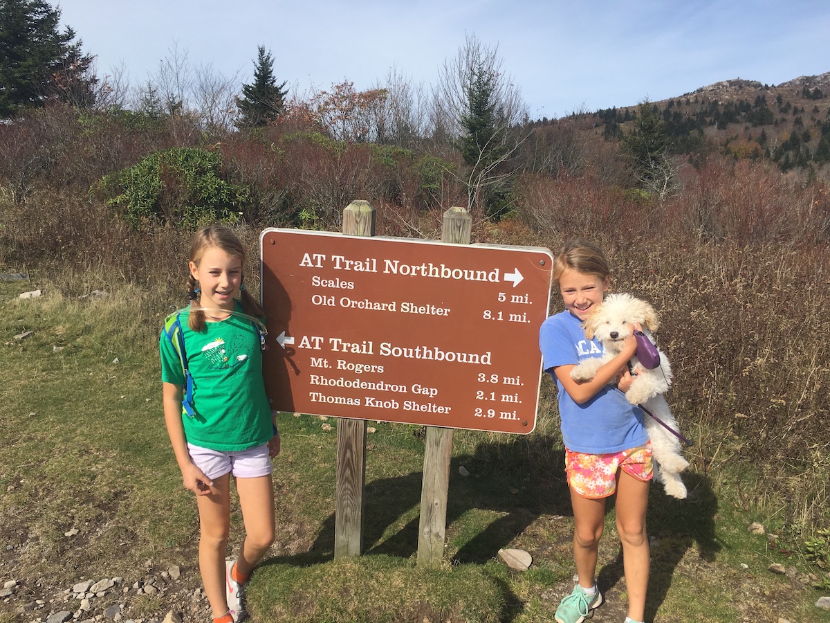 Andrew Bobilya's daughters smile next to a trailhead on a hike with their dog