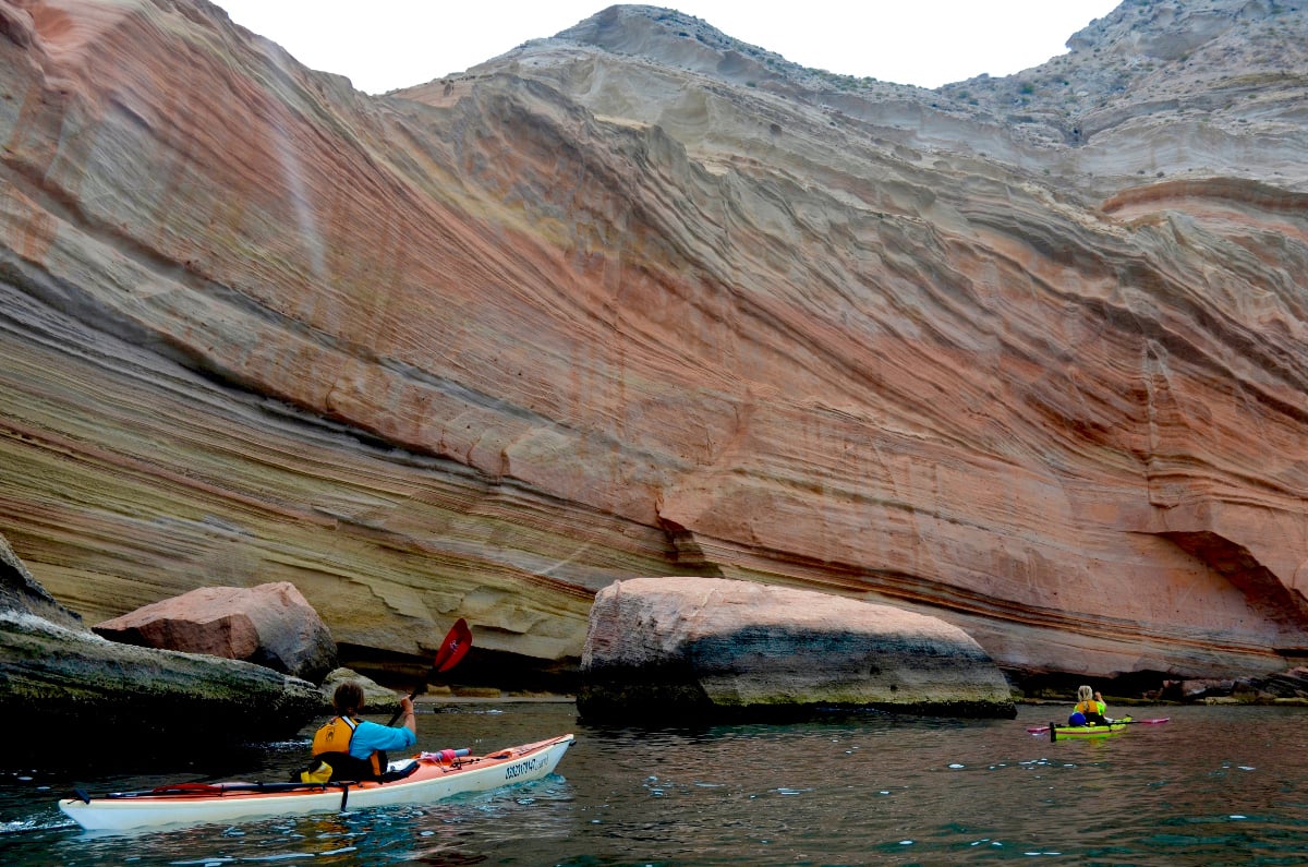 Sea kayakers paddle near wavy red sandstone wall