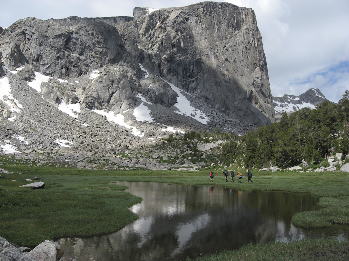 five NOLS participants hike past glassy alpine lake toward towering rocky peak