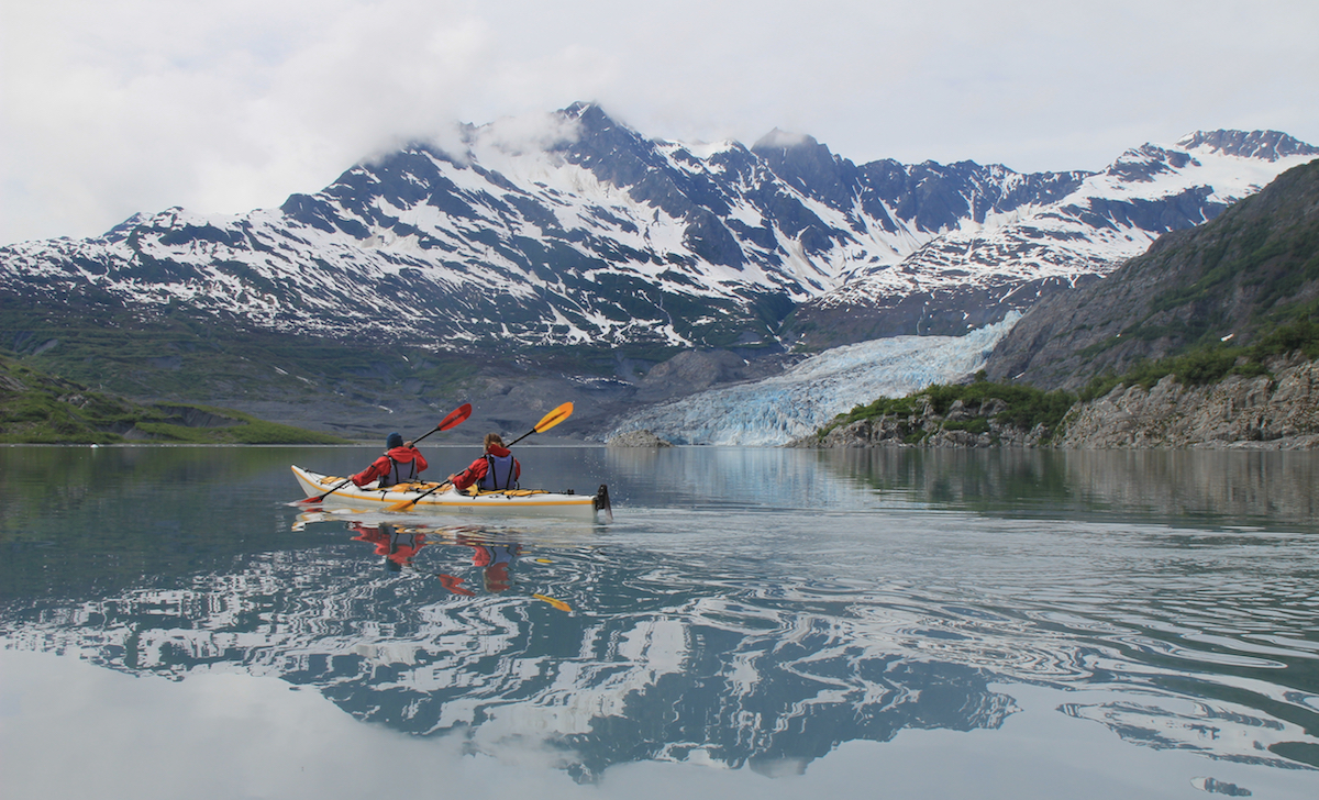 NOLS students paddle a double kayak in Prince William Sound with mountains reflected in the water