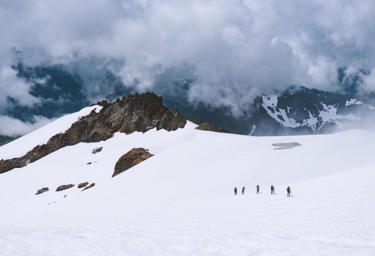 five small figures travel across a snowfield in a dramatic mountain landscape