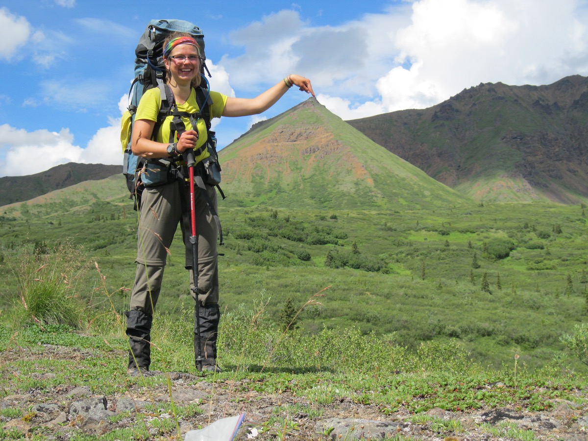 smiling female hiker wearing backpack and rainbow headband points her finger at the top of a distant mountain, creating an optical illusion