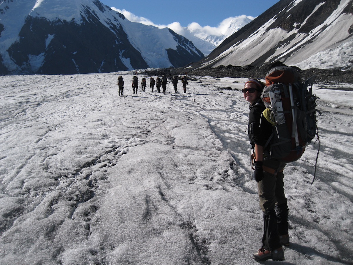 smiling NOLS student wearing backpack smiles for the camera on a glacier in Alaska with other students in the distance