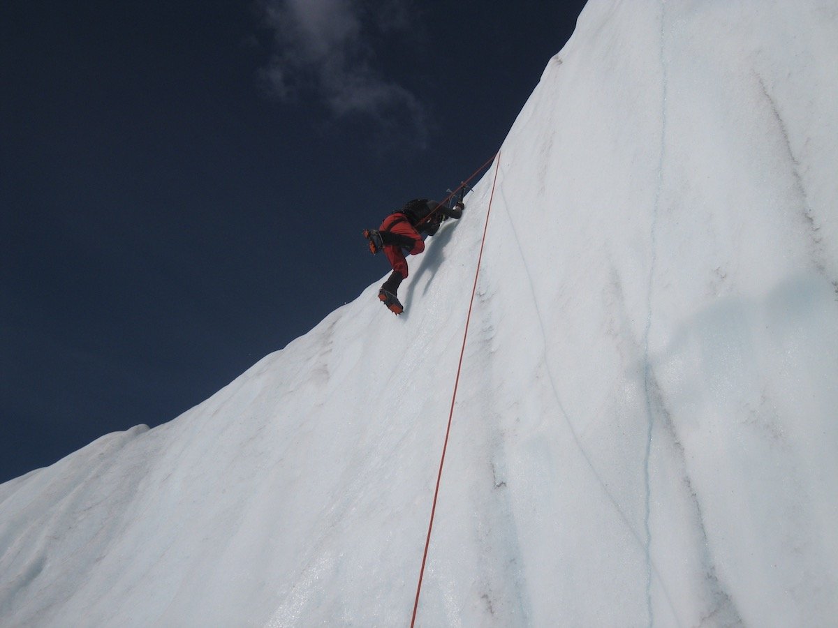 person uses crampons and rope to climb up an ice wall while mountaineering