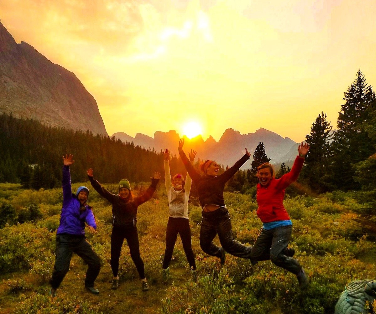 five female NOLS students smile and jump in the air while backpacking in the Rockies