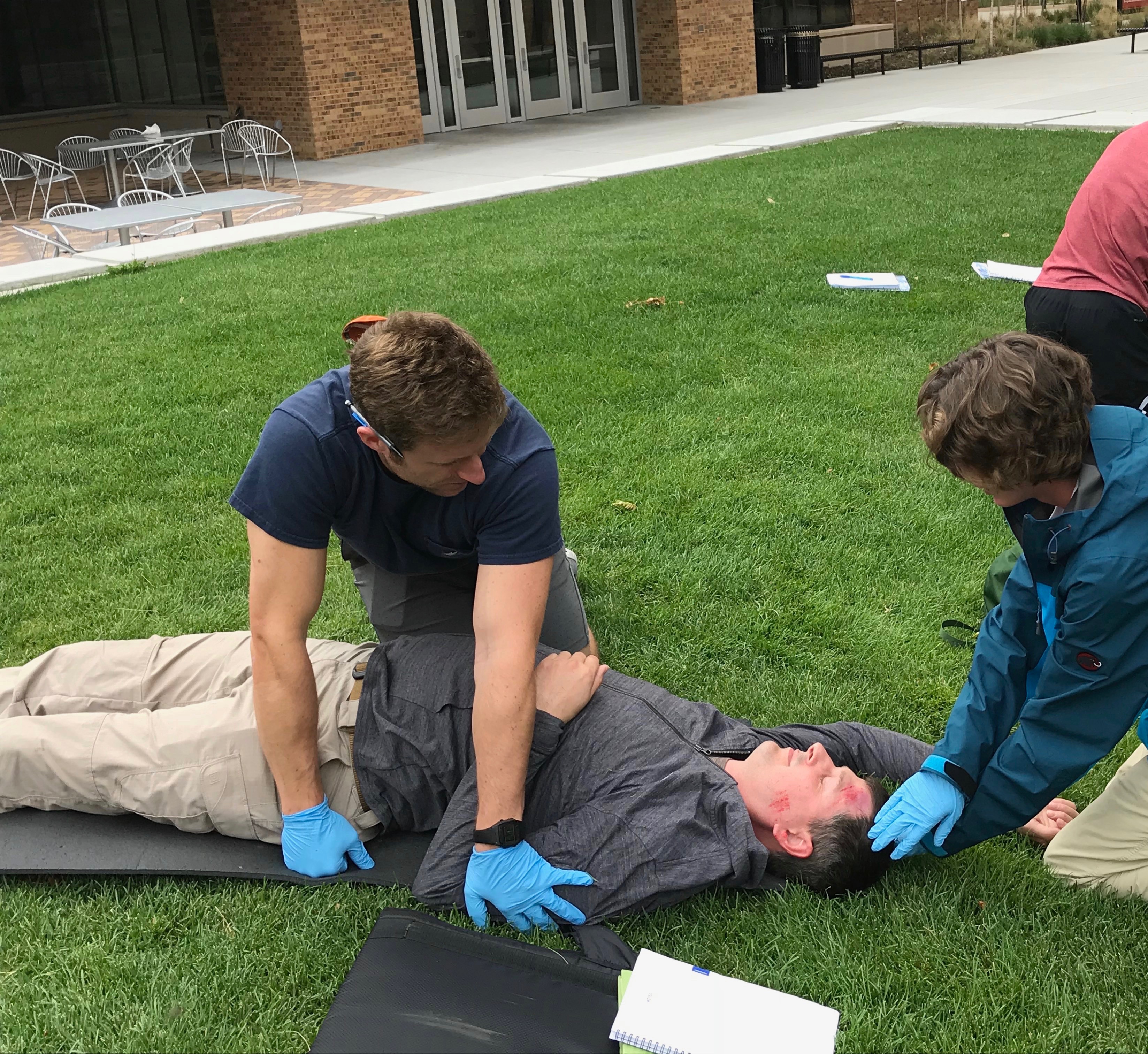 Two students, on a lawn, practice assessing a patient who is on the ground with blood on his head