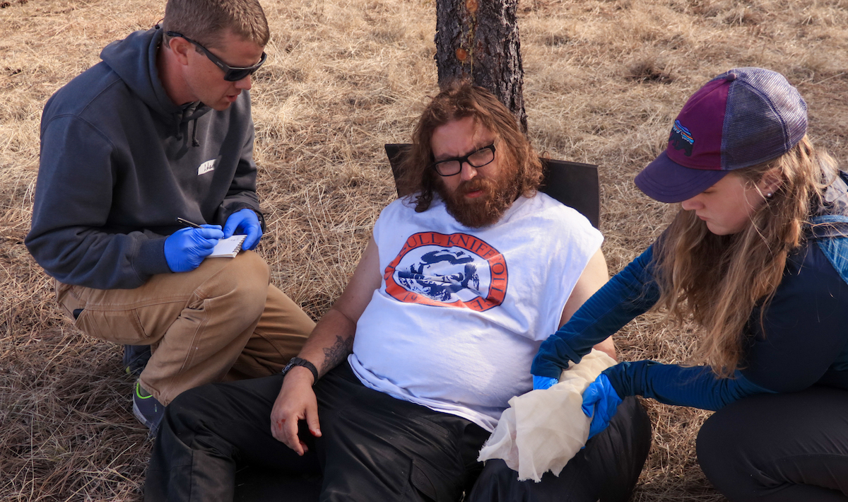 NOLS wilderness medicine students practice caring for a patient, with one writing a SOAP note and the other dressing a wound