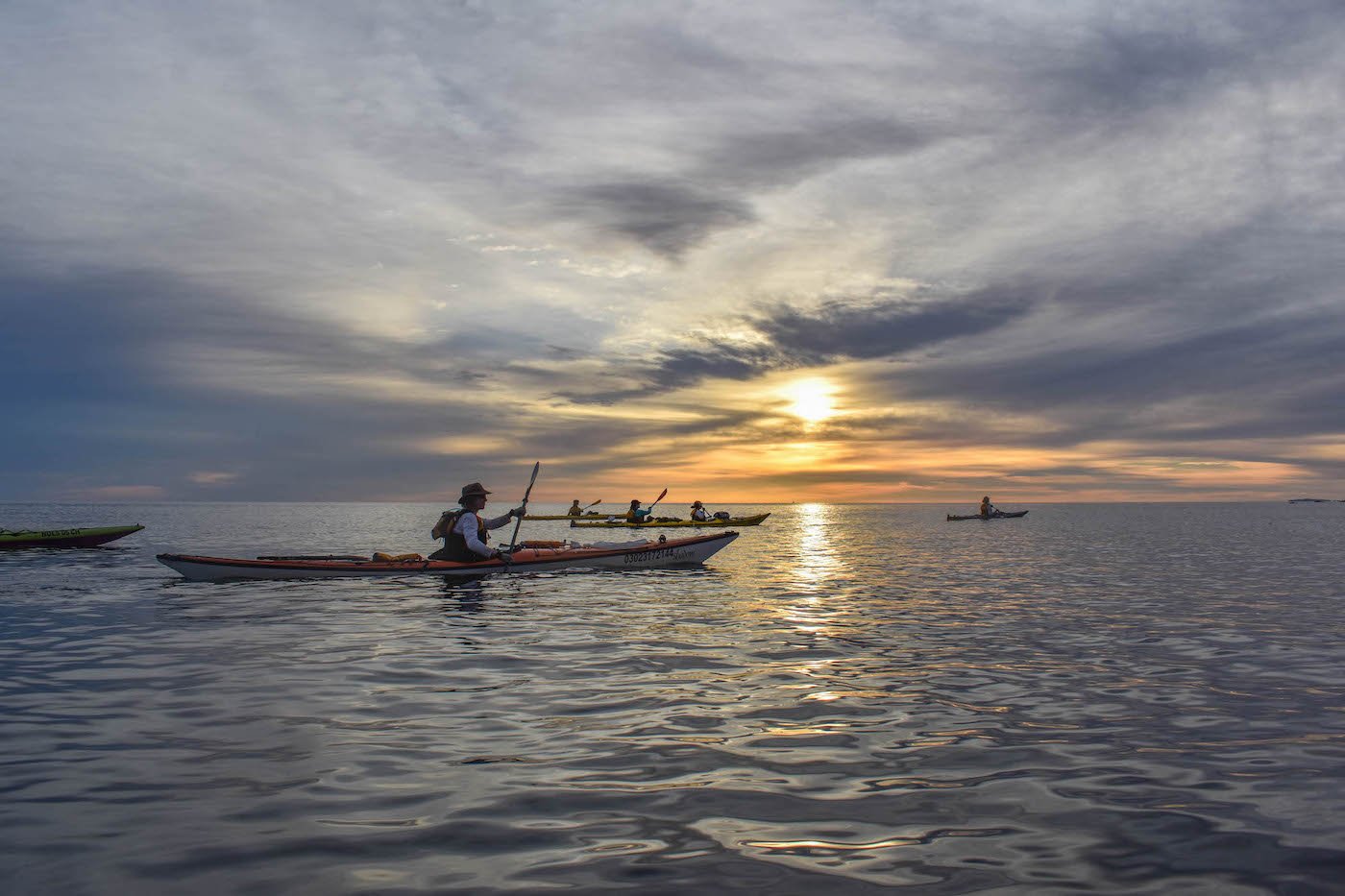 NOLS participants paddle kayaks on smooth gray water as the sun sets into the clouds over the ocean