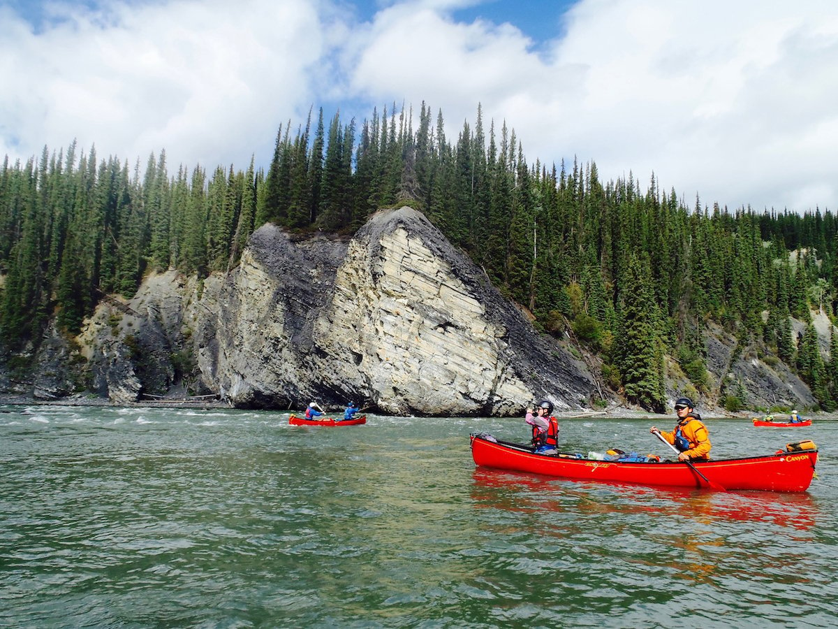 NOLS students paddle red canoes past rocky tree-lined coast in the Yukon