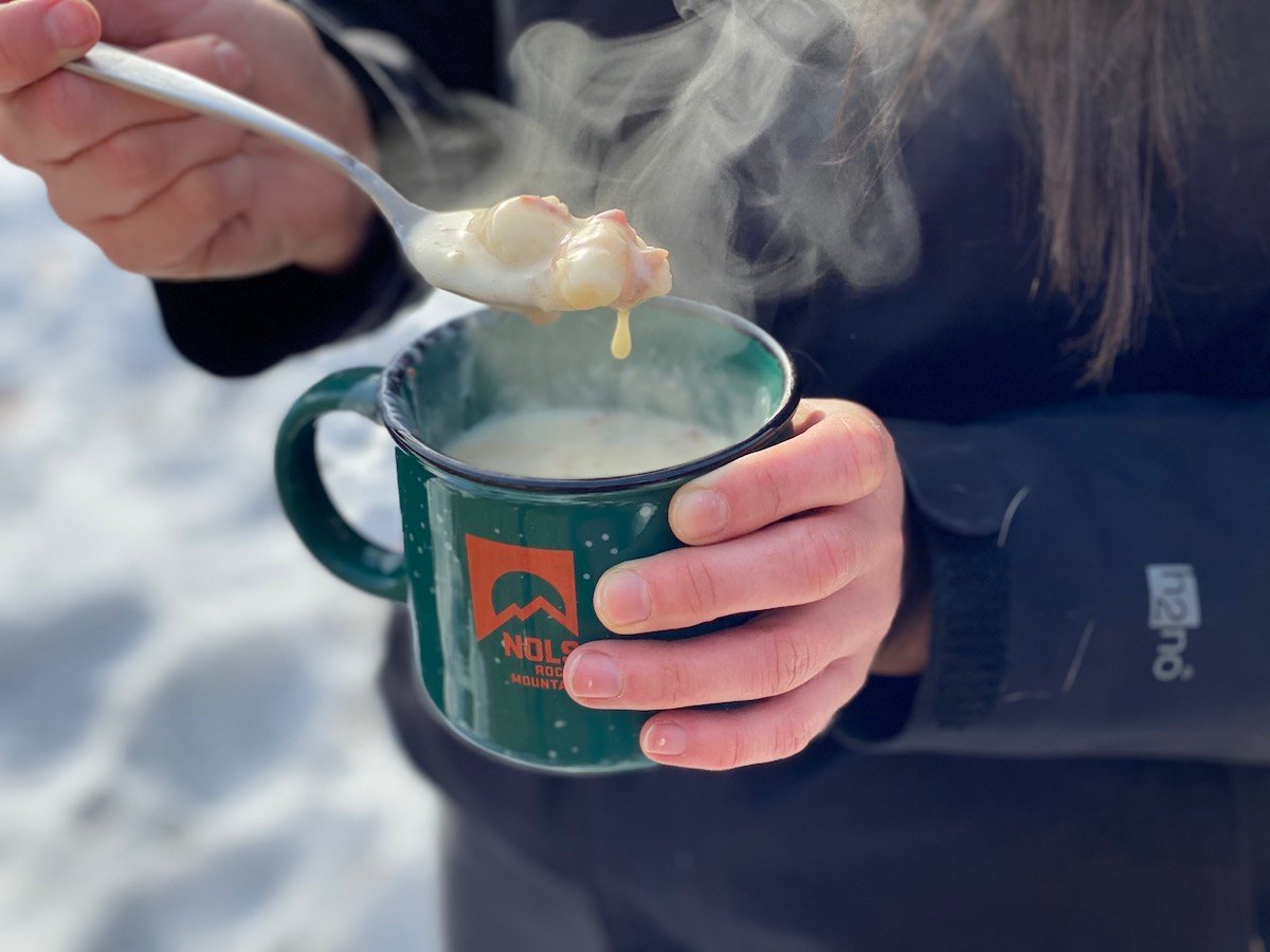 hands holding a steaming spoonful of cheesy potato soup in a green and orange NOLS mug