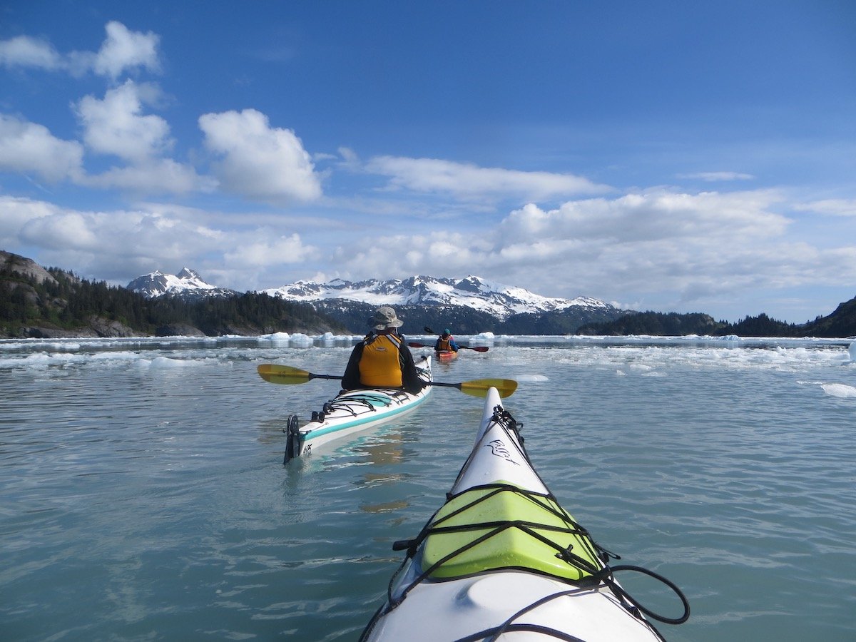 NOLS students sea kayaking in icy water with snow-capped mountains in the distance, as seen over the bow of a green and white kayak