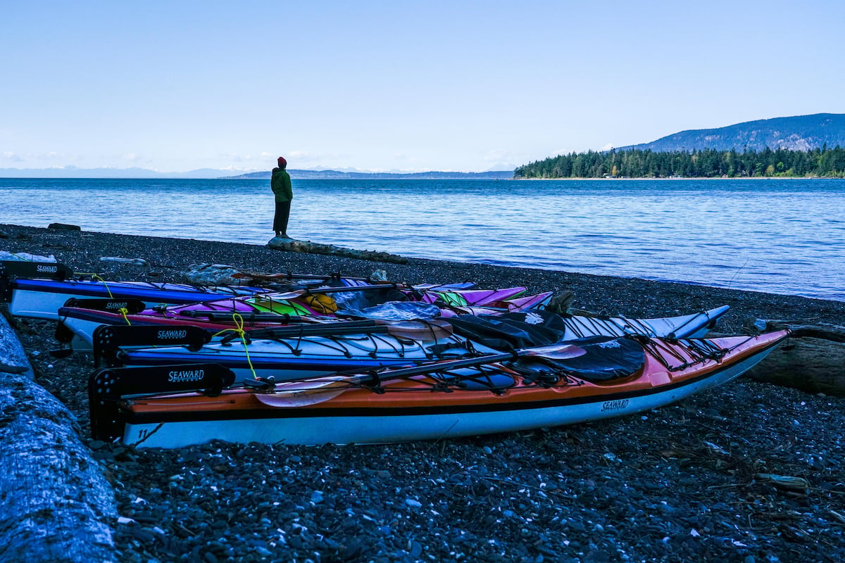 brightly colored sea kayaks on pebbly beach with lone figure gazing out at calm water