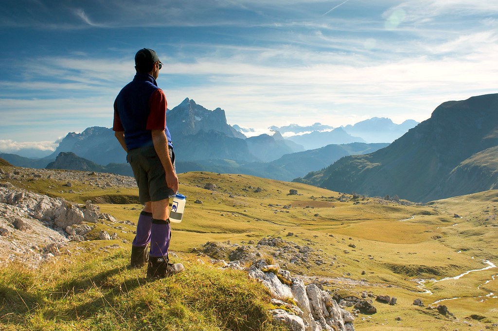 man wearing hiking gear and holding water bottle looks out over a green valley toward rugged mountains