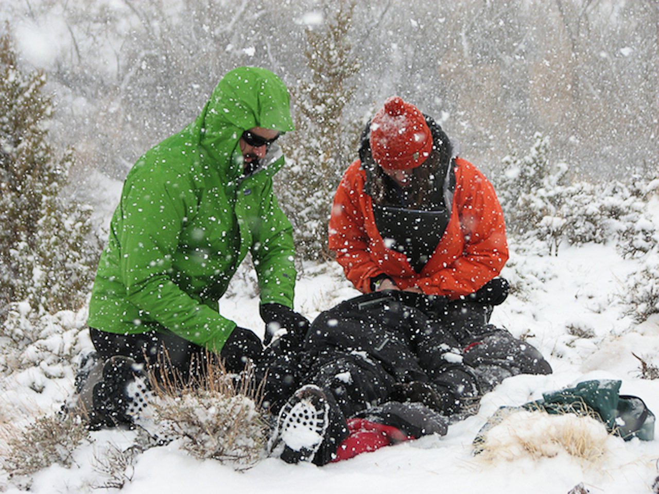 two NOLS Wilderness Medicine students practice for a patient lying on the ground on a snowy day