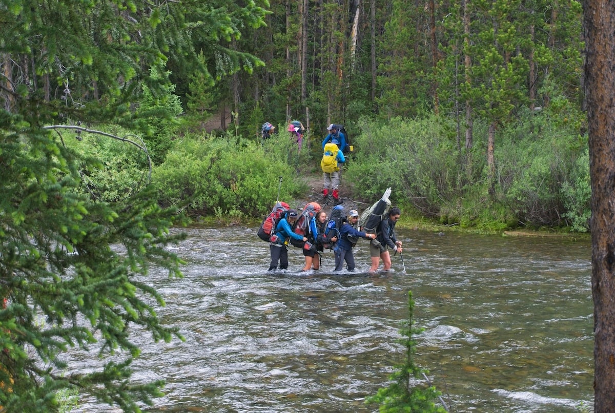 students walk down a trail to the river as other students wearing backpacks cross a river surrounded by thick forest