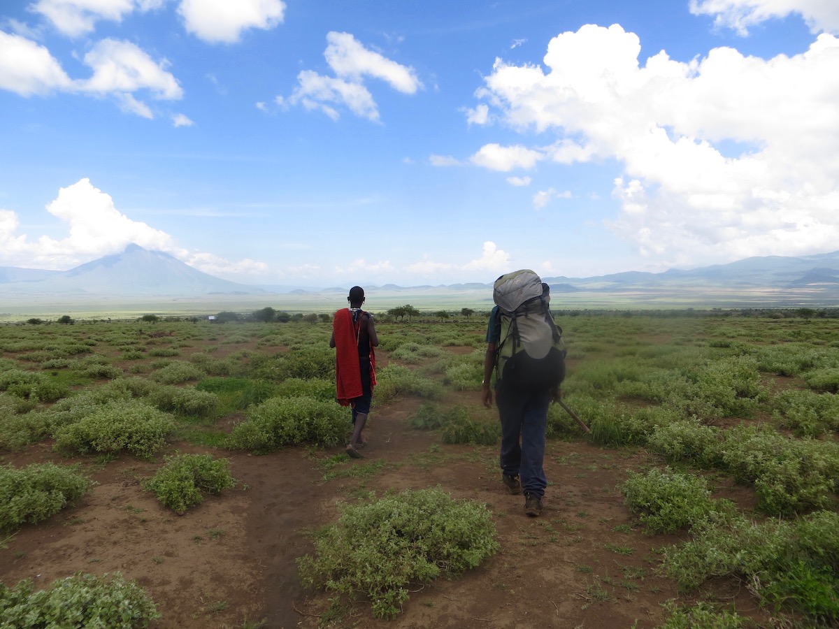 Two people, seen from behind, backpack through Maasai Land in East Africa with the slopes of Mt Kilimanjaro in the distance