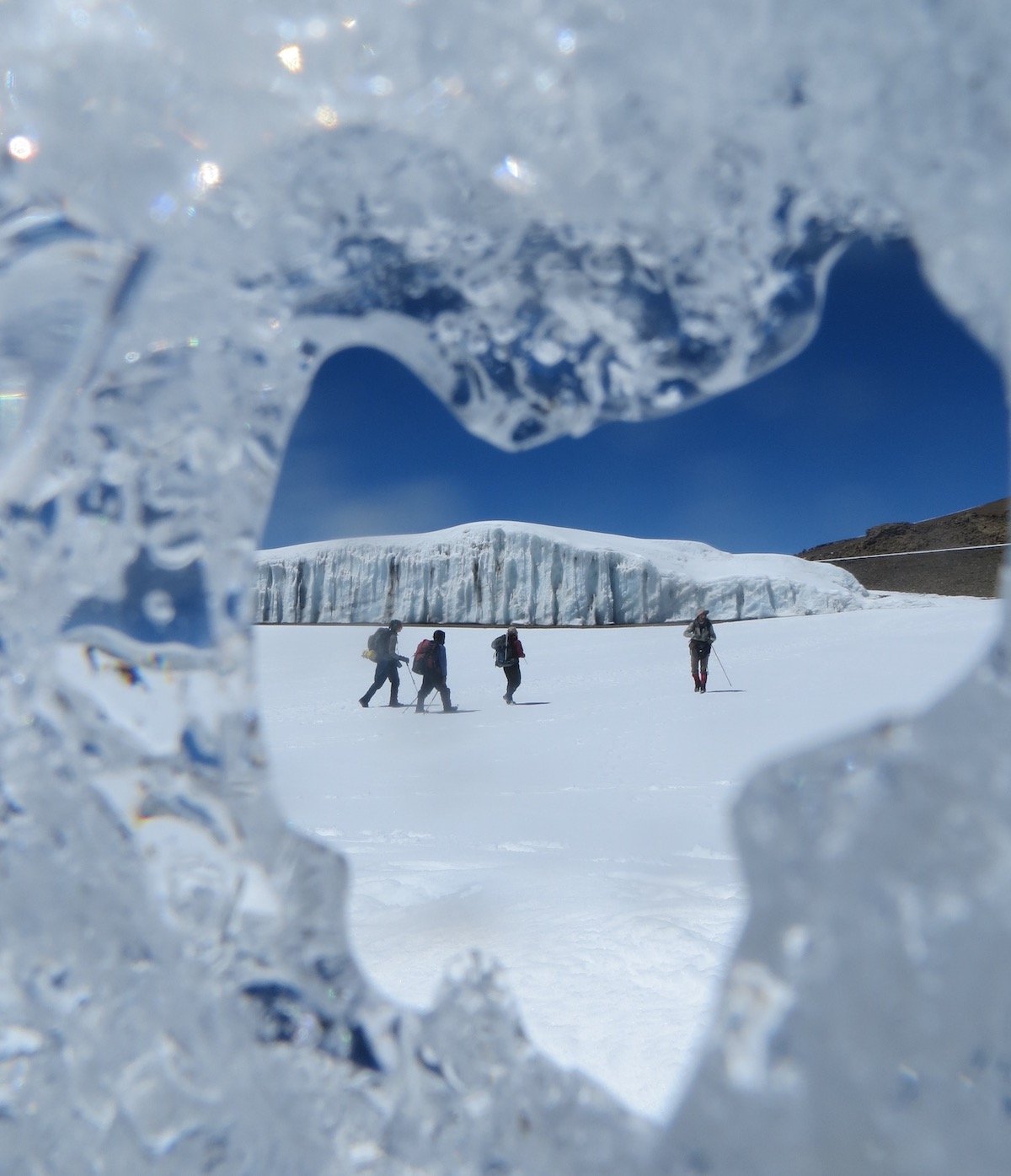 four students hike across a snowfield on Mt Kilimanjaro, as viewed through a crack in a melting glacier
