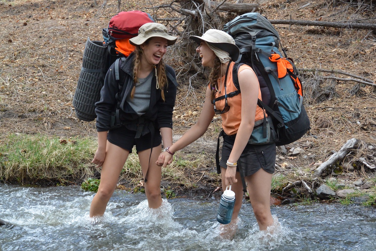 two smiling teenage girls wearing backpacks hold hands while crossing a river