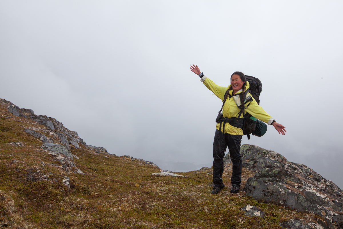 smiling woman with backpack stands on rocky ground and flings up her arms on a foggy day in the mountains