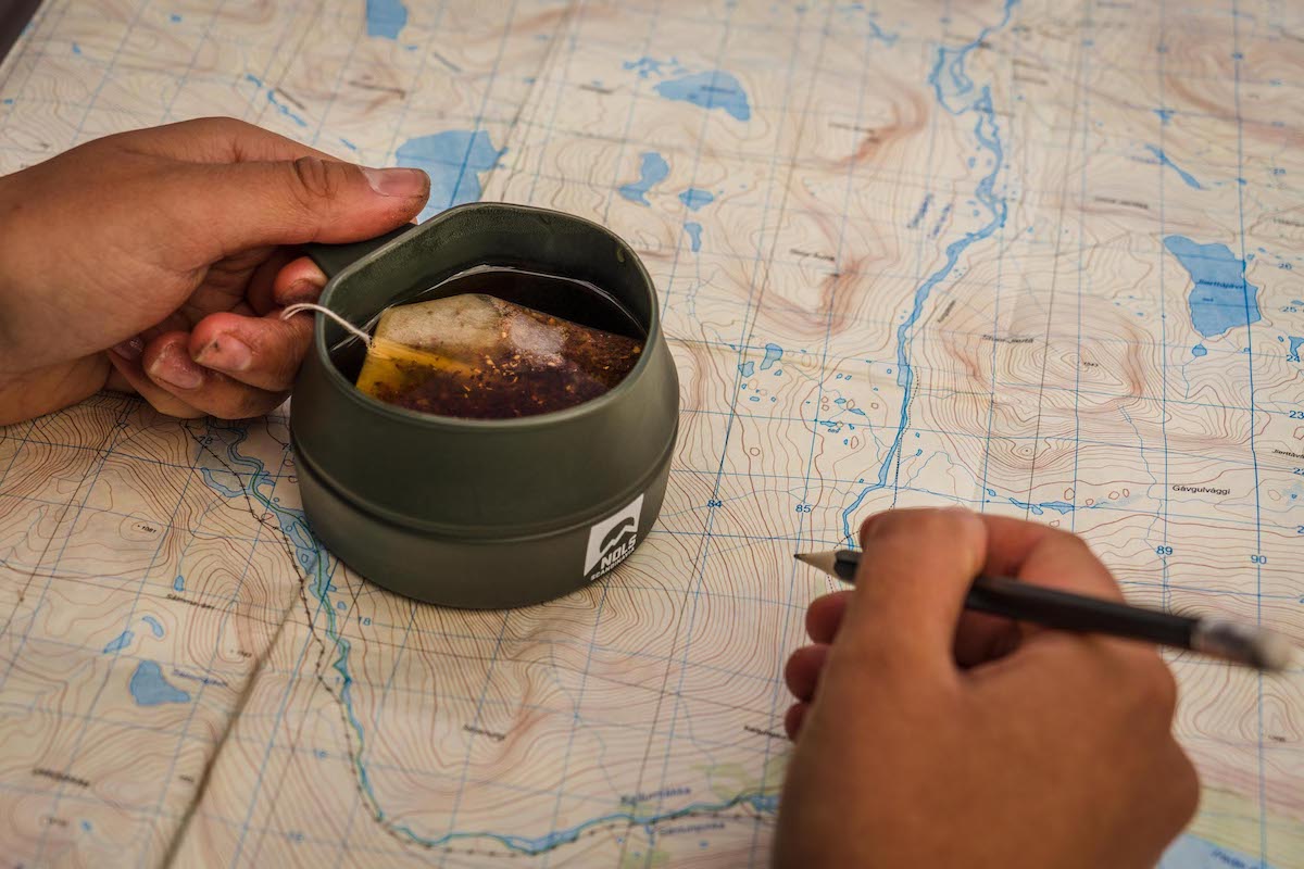 two hands on a topo map with the left hand holding a mug of tea and the right holding a pencil