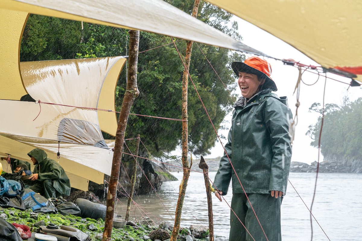 woman wearing orange hat and rain gear laughs while standing under a tarp near the water's edge in a rain storm