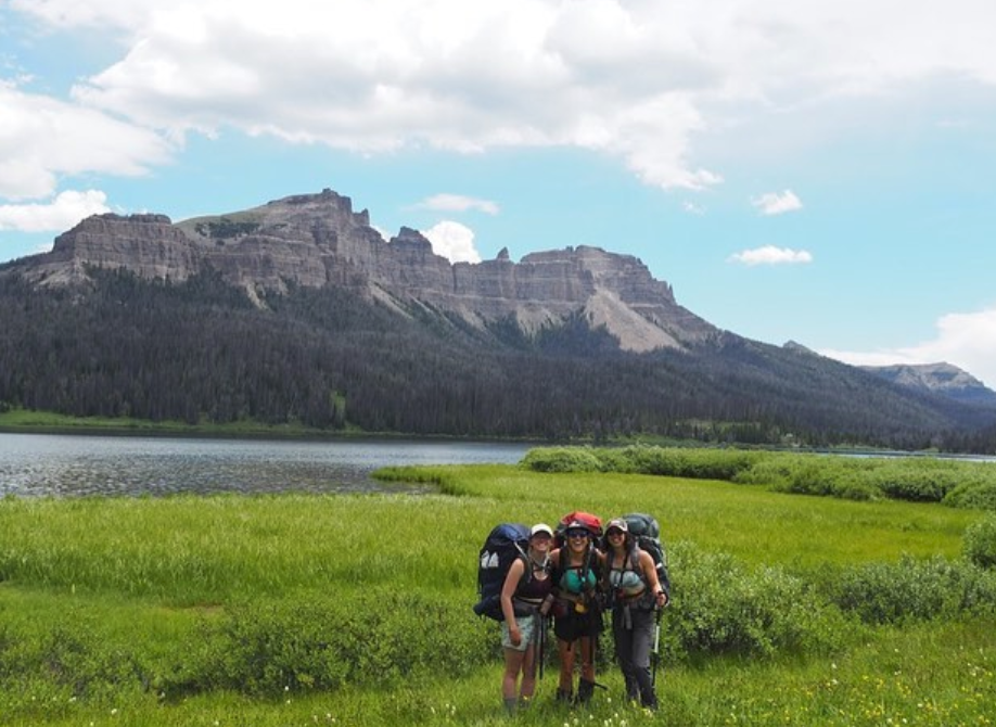 three smiling female students stand in a green meadow near a lake with rocky mountains behind
