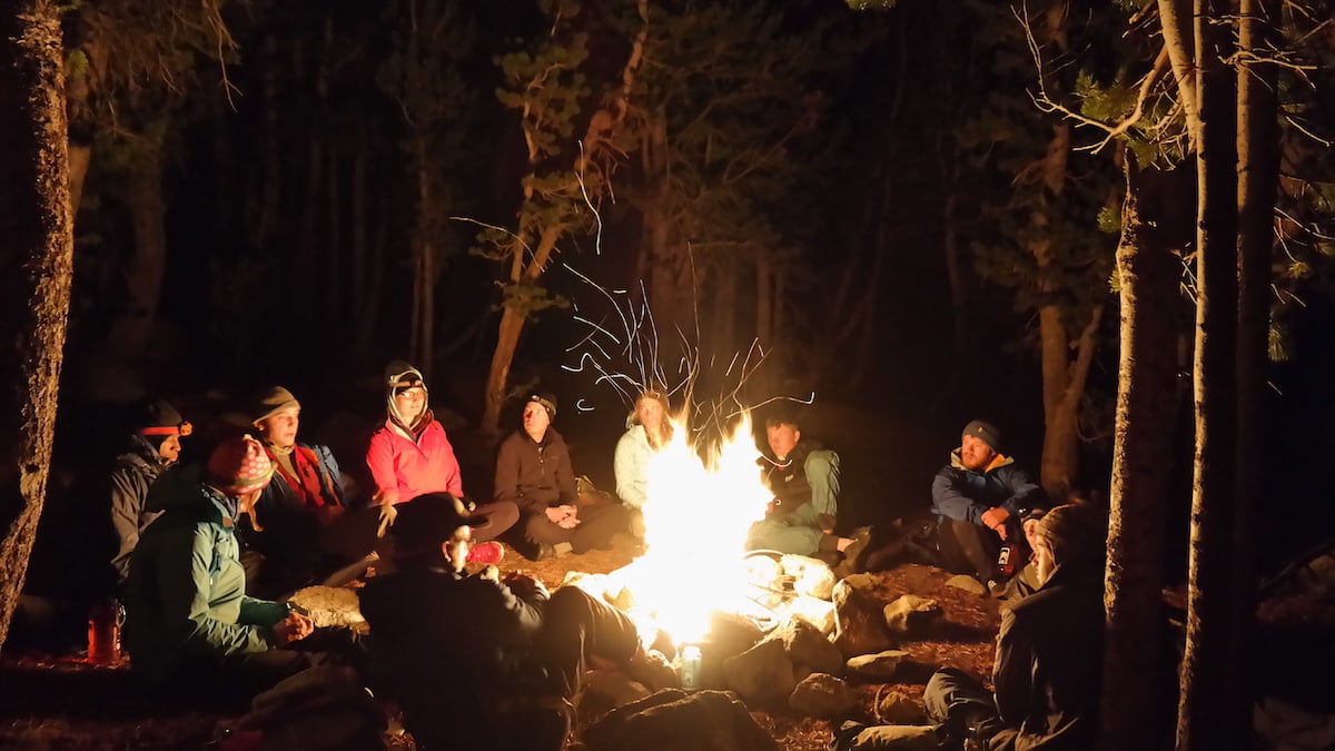 group of people gather around a campfire in the woods at night
