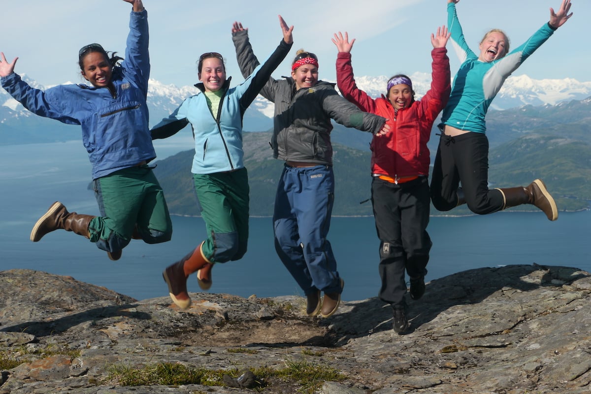 five female NOLS students smile and leap into the air on a rocky ledge in Alaska