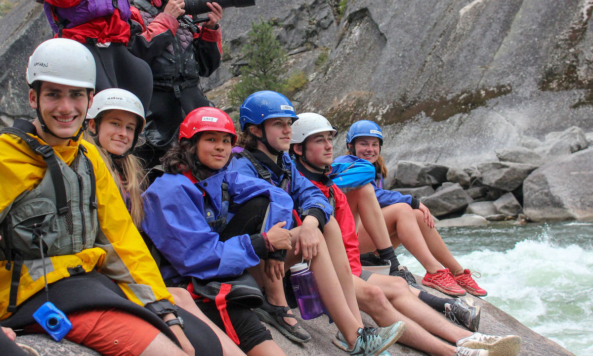 six teenage NOLS students sit on a rock wearing helmets on a Salmon River rafting course