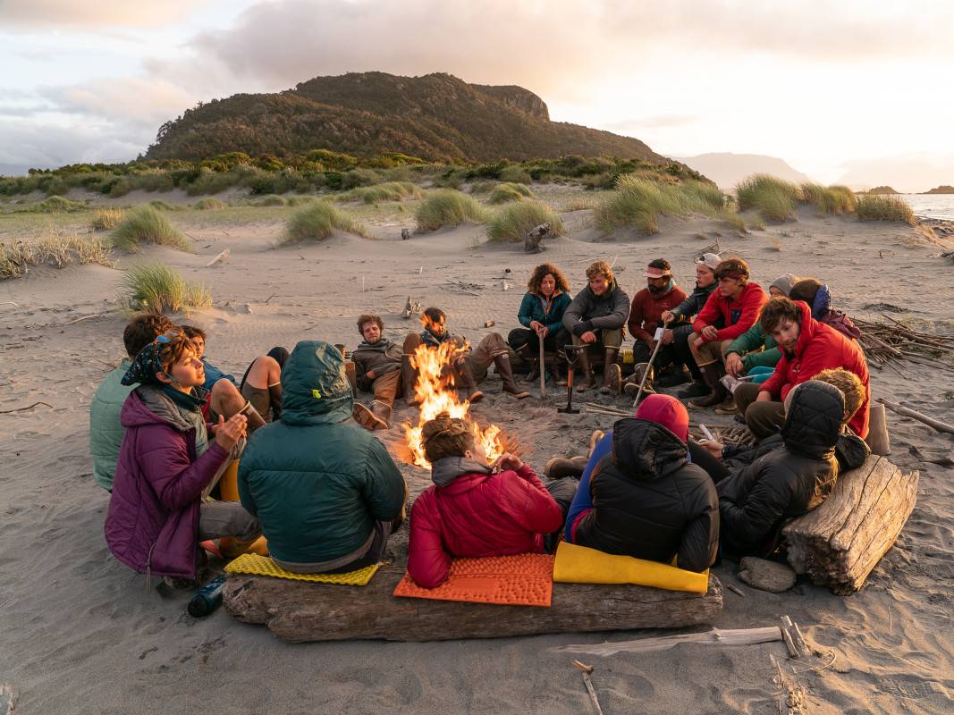 Group sits in a circle around a fire on a sandy beach