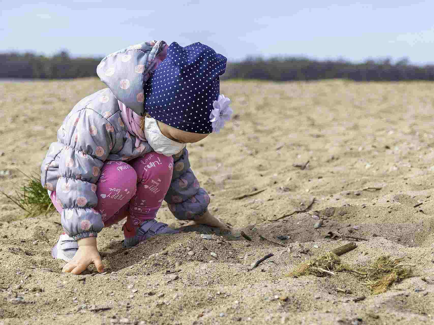 Toddler bends over the sand while wearing a hat and face covering