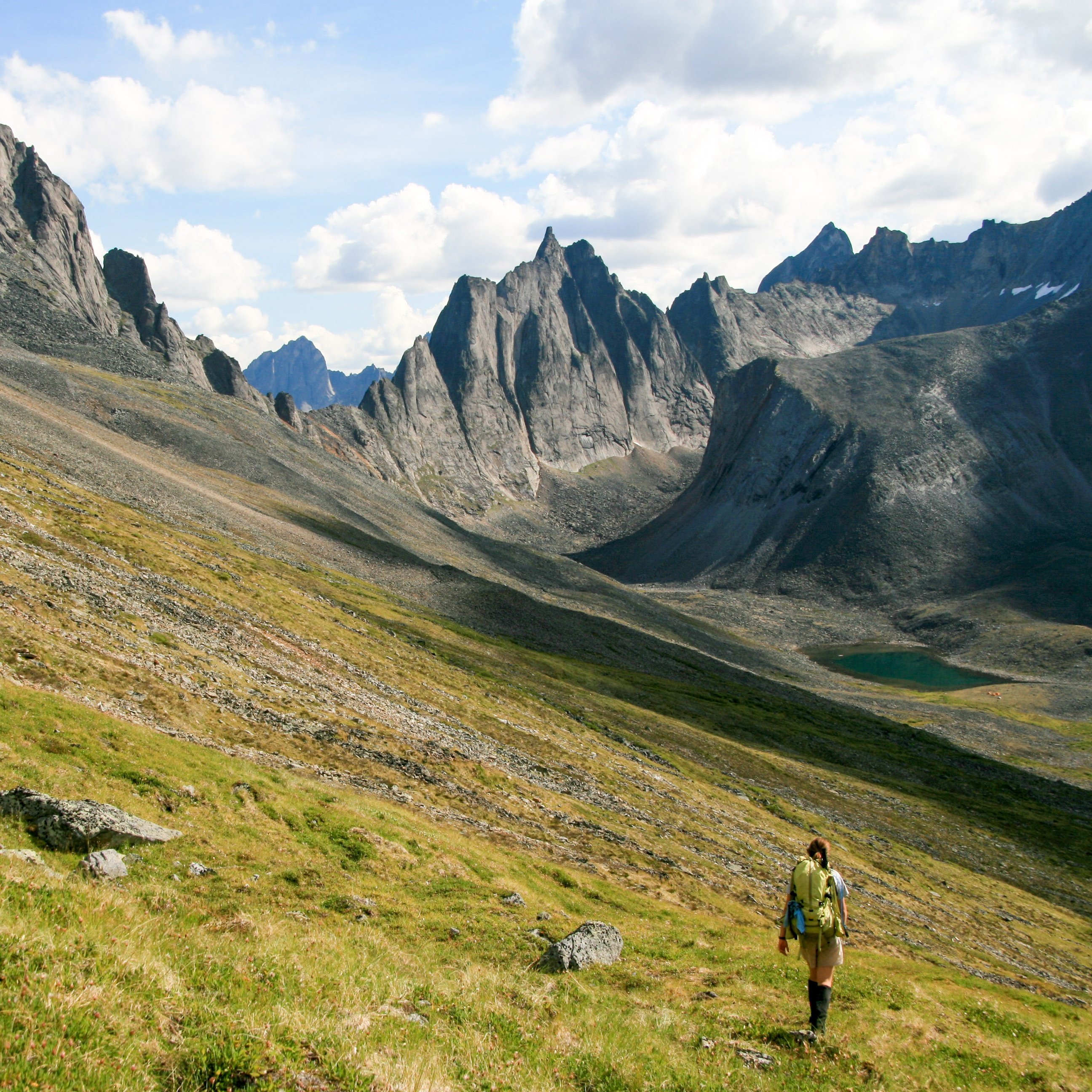 Backpacker hikes through grassy foothills in the Yukon with high, steep peaks in the background