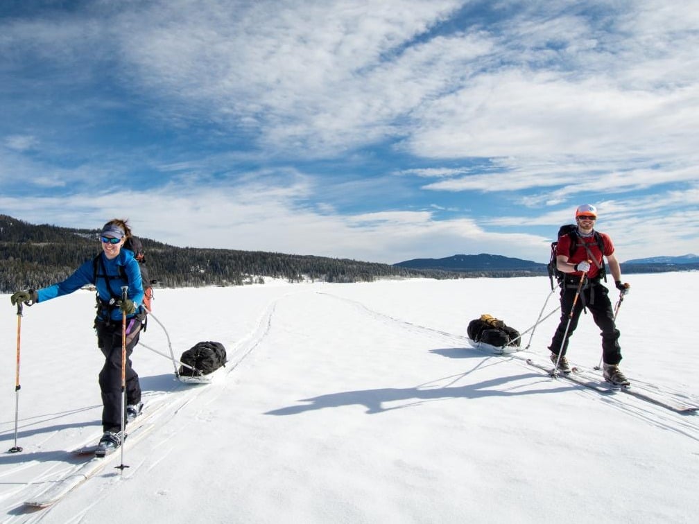 Two smiling people ski touring and pulling sleds across a snowfield