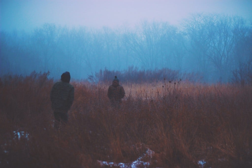 two people walk in tall scrub brush with patchy snow in the cold