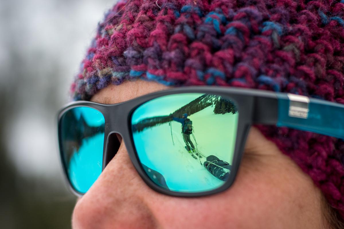 Close-up of person wearing sunglasses and knit hat with person on skis pulling sled reflected in the glasses