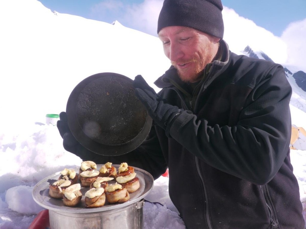 Man lifts the lid on a pan of cinnamon rolls in a snow kitchen