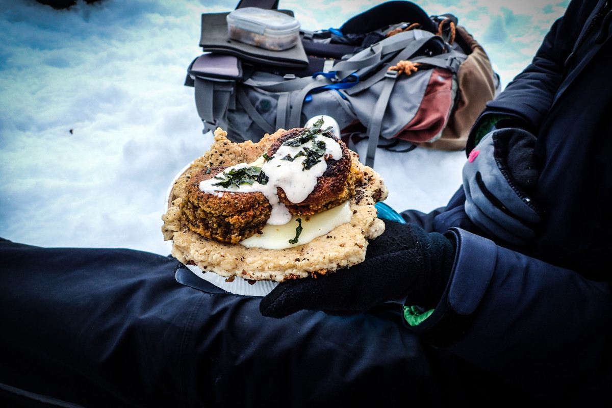 Person wearing gloves and winter gear sits in the snow holding a serving of backcountry falafel with a backpack nearby