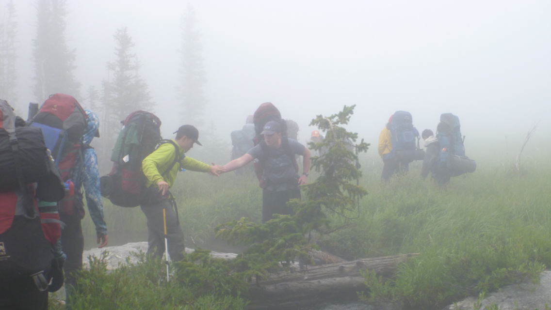 A group of backpackers cross a foggy field in the Bighorn Mountain Range in Wyoming. One boy helps another across a log.