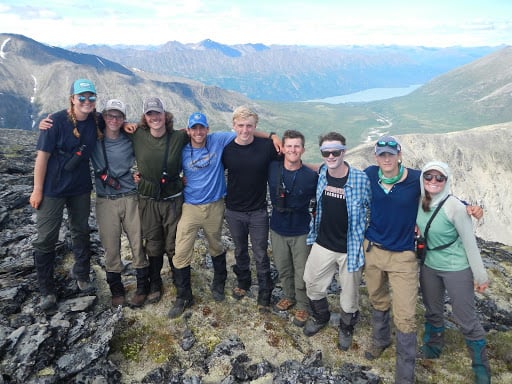 Nine smiling NOLS students stand in a line with arms around each other in Alaska's mountains
