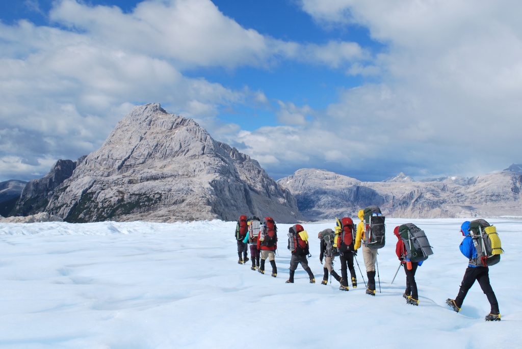 line of NOLS students hike across a snowfield toward rocky peaks using trekking poles