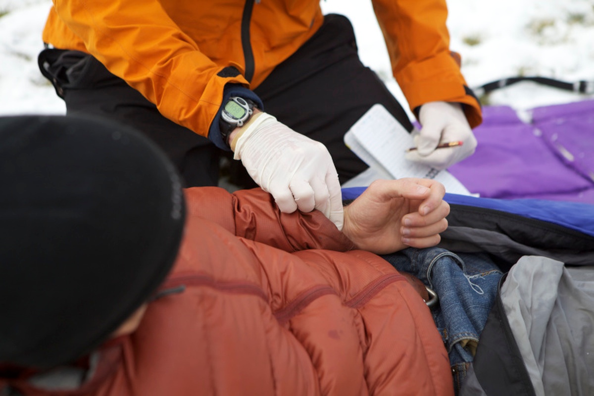 Person wearing orange jacket and white rubber gloves practices taking vitals on a patient lying in the snow